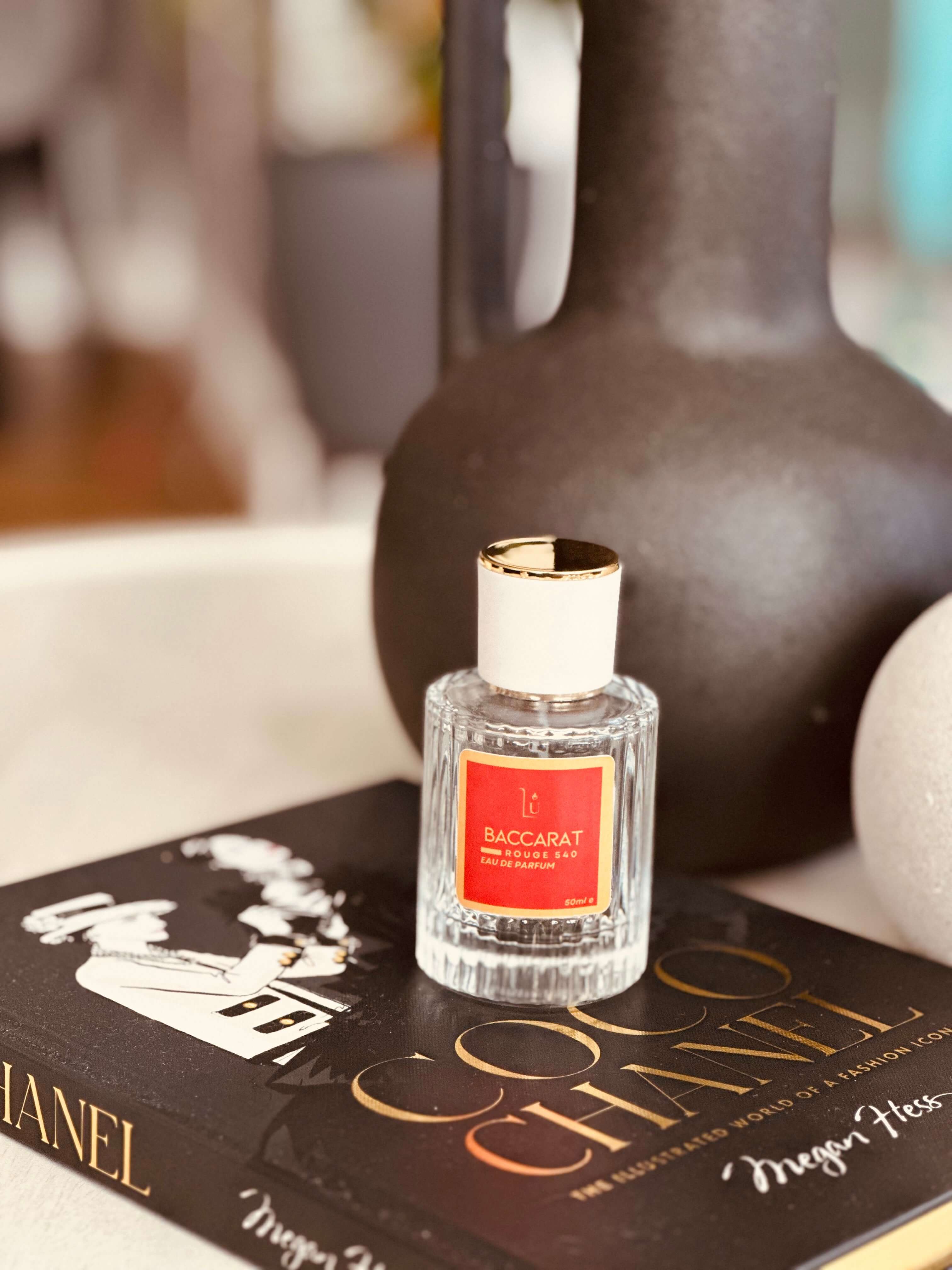Amber & Saffron Eau de Parfum by LU Fragrances — 50ml glass bottle with red label and white gold cap, displayed on a Coco Chanel book with a large dark matte ceramic vase and cream sphere in the background
