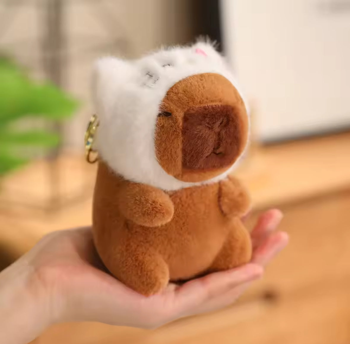 Plush toy resembling a capybara with a white cat hat held in a hand on a wooden surface.