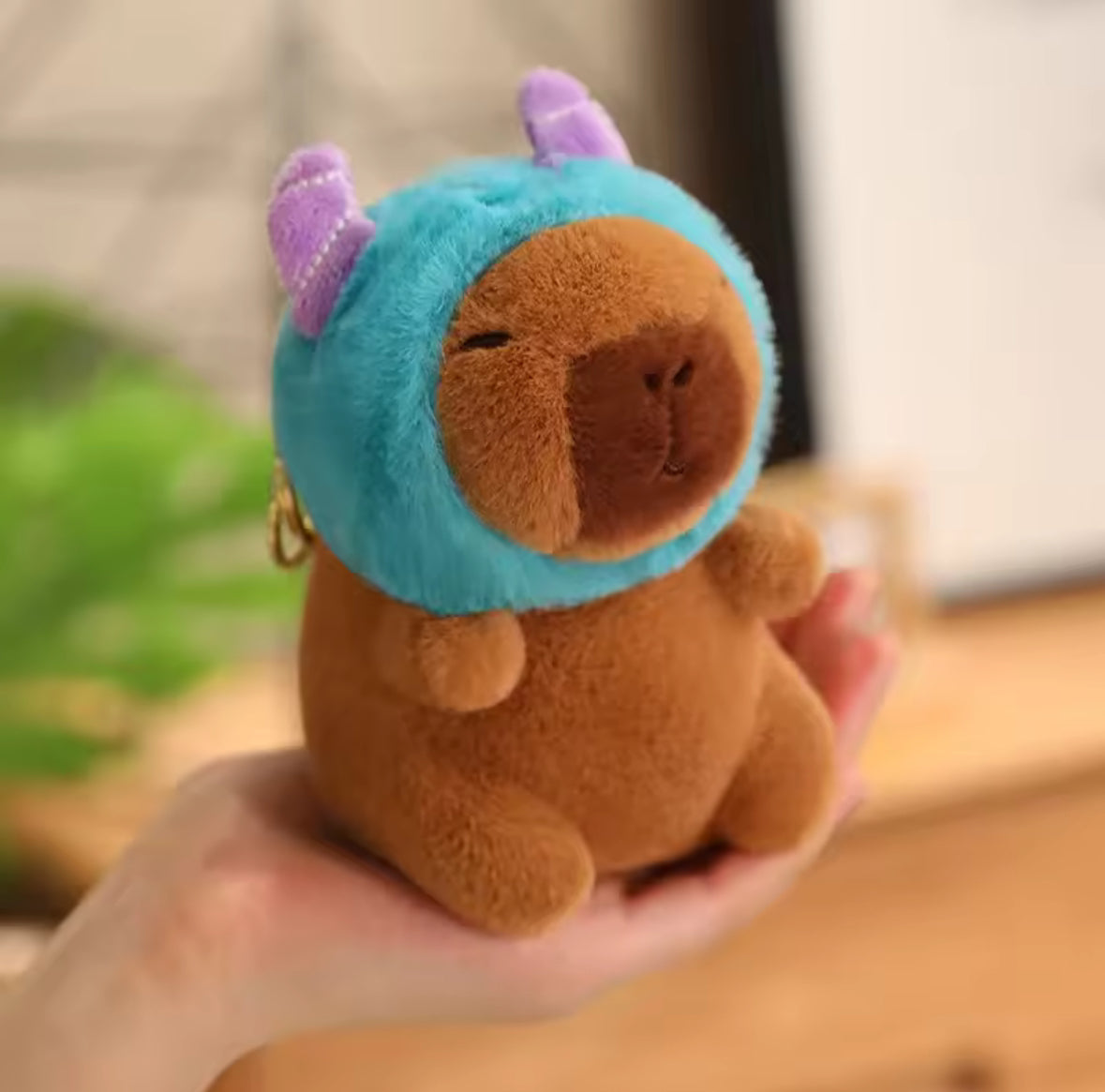 Brown plush capybara with a blue hood and purple accents held in a hand against a blurred background