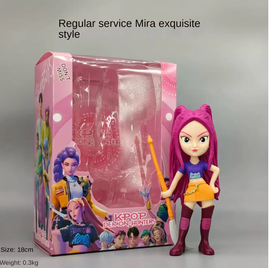 K-pop doll with pink hair and colourful outfit standing next to its packaging on a grey background