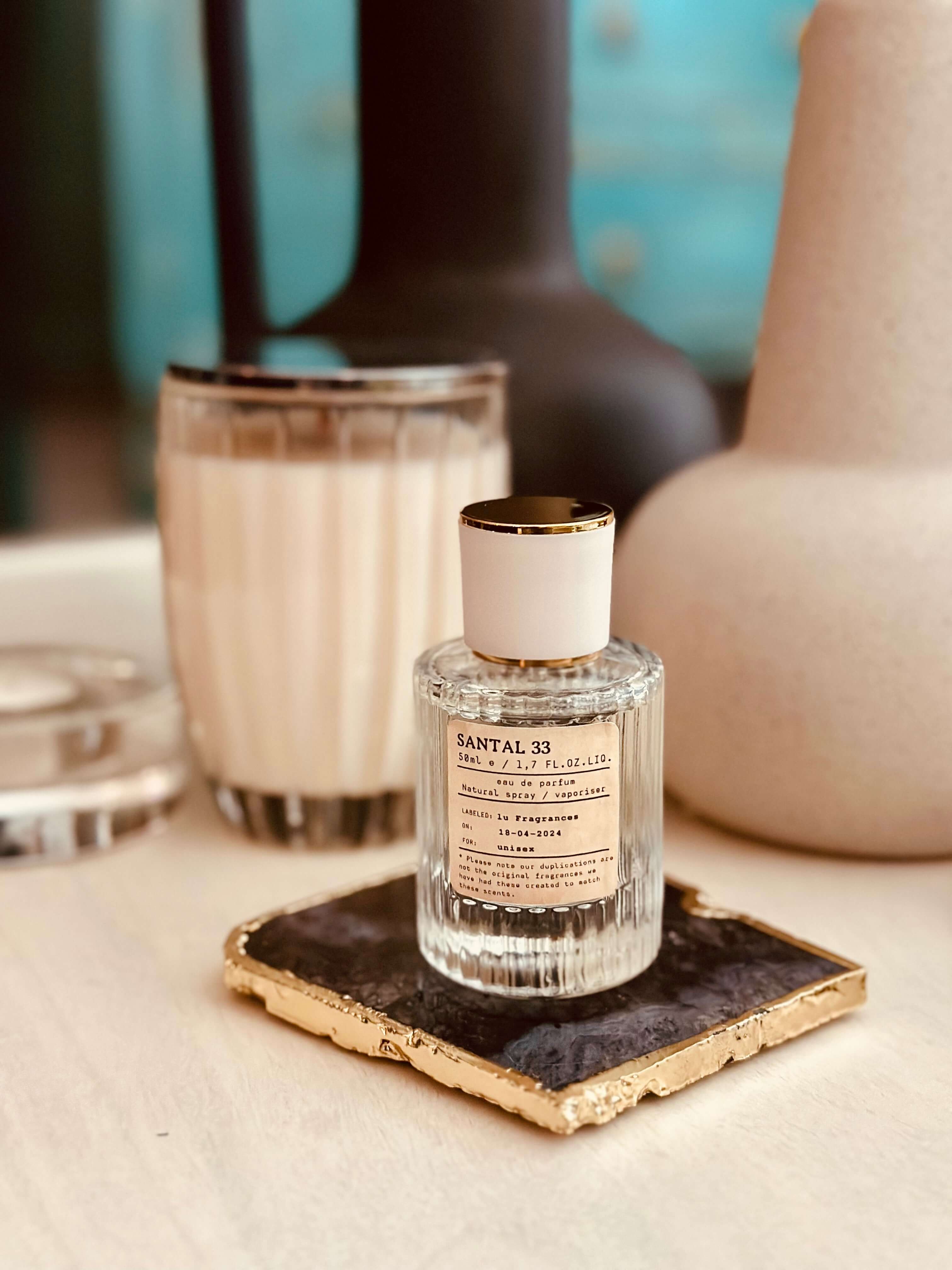 Sandalwood & Cardamom Eau de Parfum by LU Fragrances — 50ml glass bottle with cream label and white gold cap, displayed upright on a gold-edged black stone coaster with a white candle and dark ceramic vase in the background