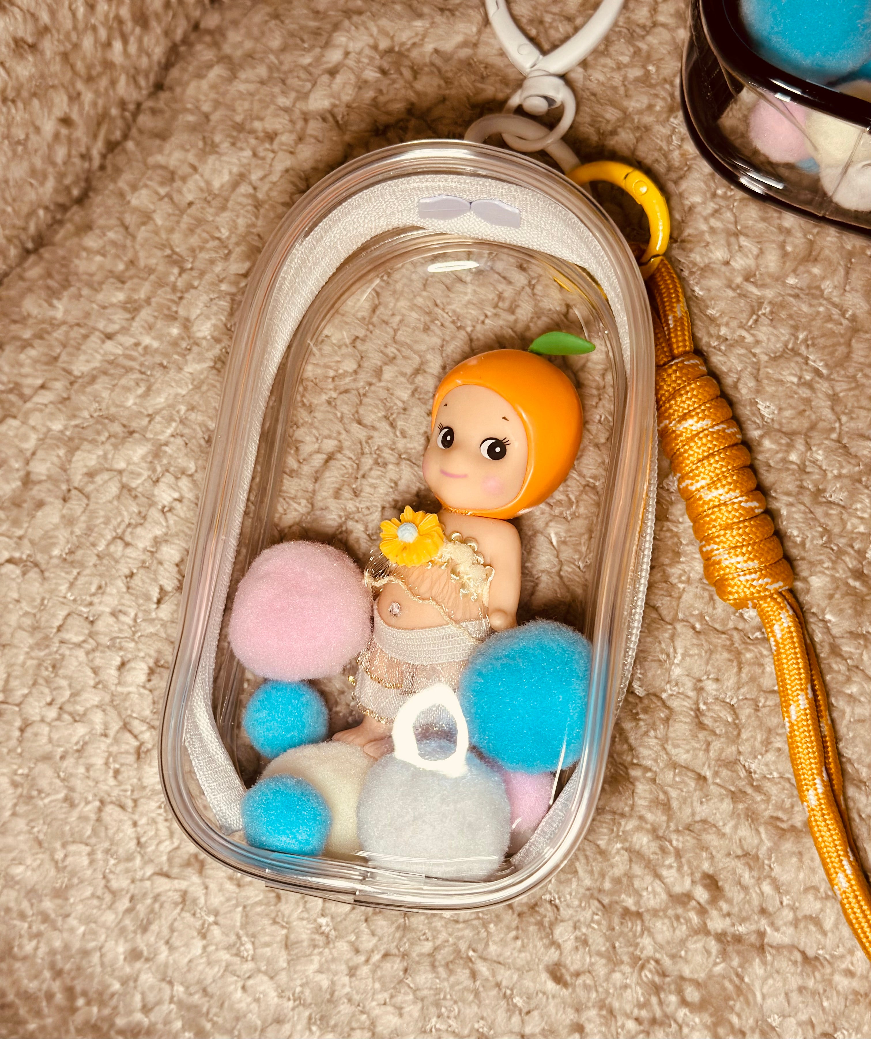 Small figurine with colorful pom-poms inside a clear container on a textured surface