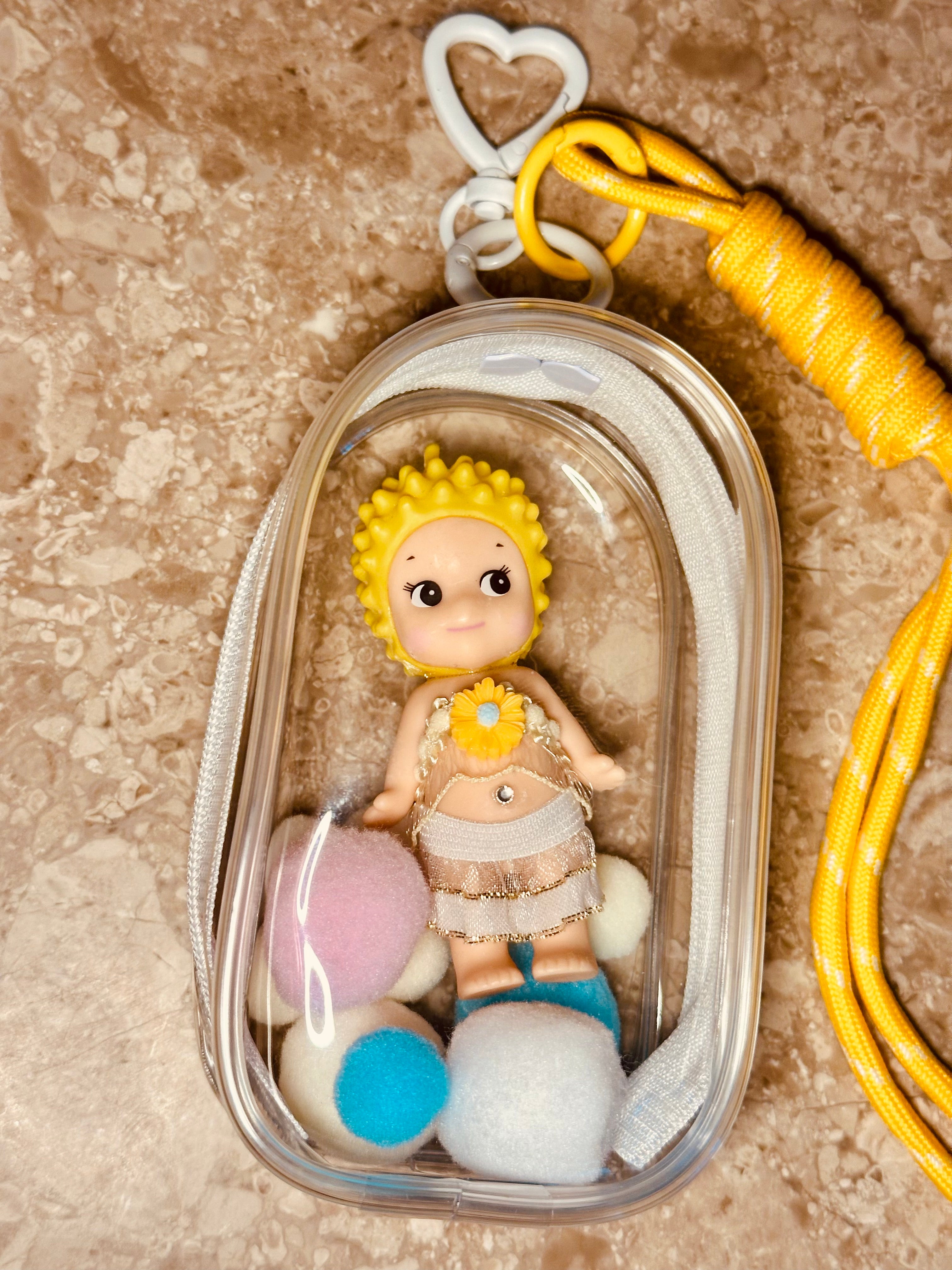 Small figurine of a baby with yellow hair and a sunflower, inside a clear plastic case on a textured surface.