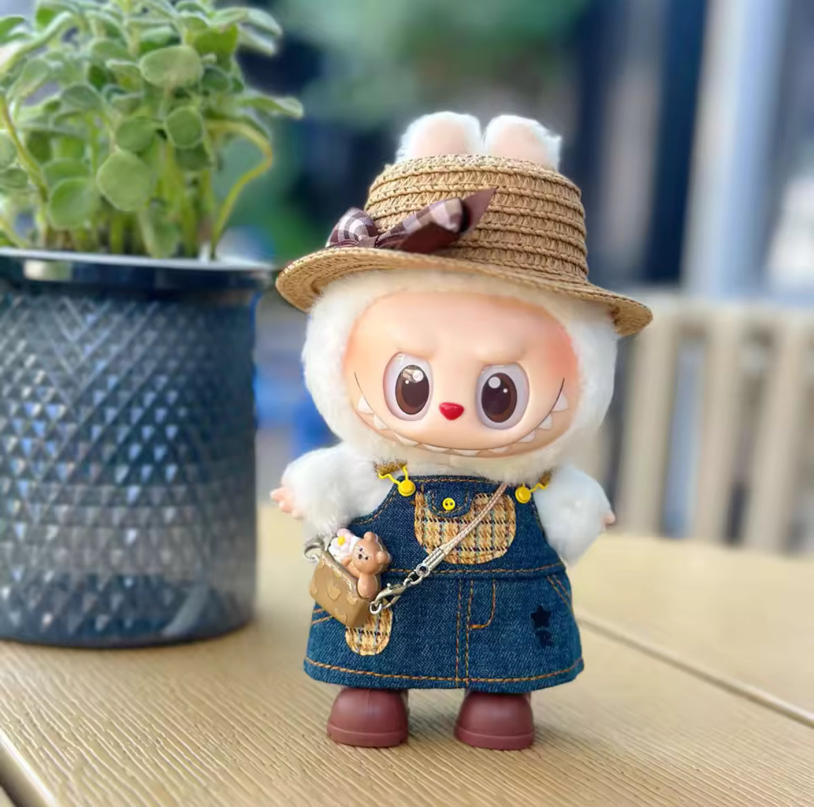 Small figurine with a straw hat and denim overalls next to a potted plant on a wooden surface.