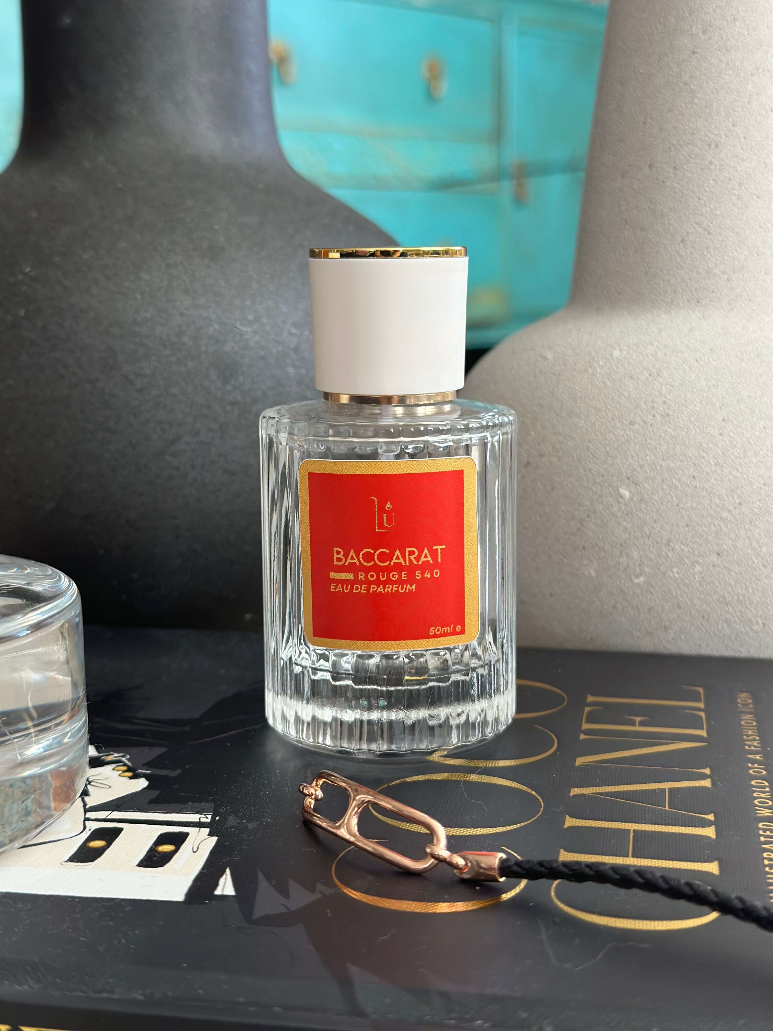 Amber & Saffron Eau de Parfum by LU Fragrances — 50ml glass bottle with bold red label and white gold cap, styled on a Coco Chanel coffee table book with a dark ceramic vase and gold chain in the foreground