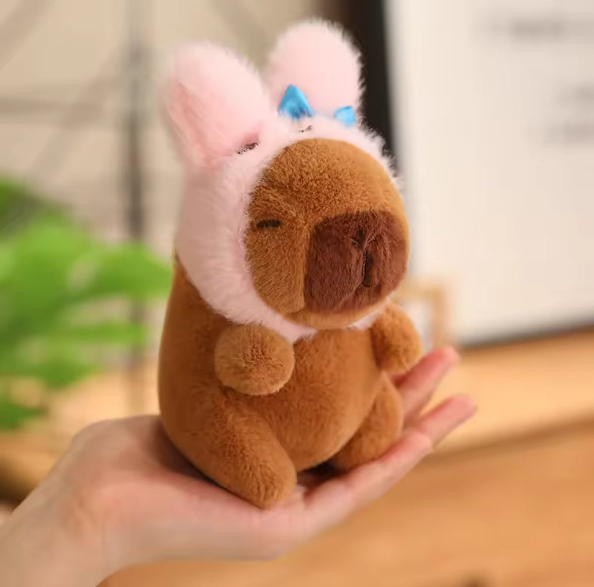 Plush toy resembling a Capybara with a pink hat with ears on it's head, held in a hand against a blurred indoor background
