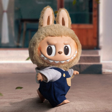 Crafted from a combination of vinyl and plush, this doll stands approximately 40cm's tall and is dressed in denim dungarees paired with a pastel blue shirt, predominantly showcasing beige and blue tones.