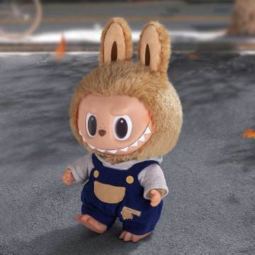 Crafted from a combination of vinyl and plush, this doll stands approximately 40cm's tall and is dressed in denim dungarees paired with a pastel blue shirt, predominantly showcasing beige and blue tones.
